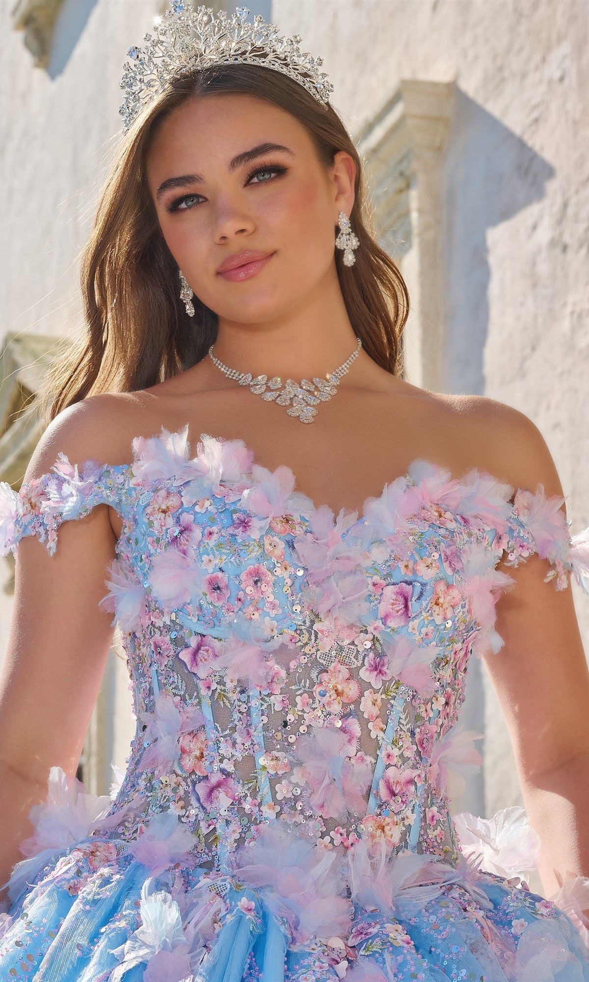 A young woman in the Quinceanera Dress 54228 by Amarra, featuring 3D floral appliqué and an off-the-shoulder design, wears a jeweled tiara and diamond jewelry while standing against a light-colored outdoor wall.